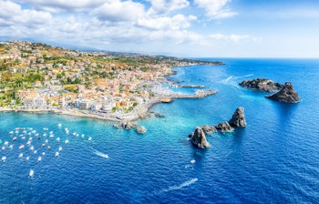 Luftfoto af Aci Trezza på Siciliens østkyst med de berømte Kyklopiske øer (Faraglioni) der rejser sig majestætisk fra det turkisblå Ioniske Hav