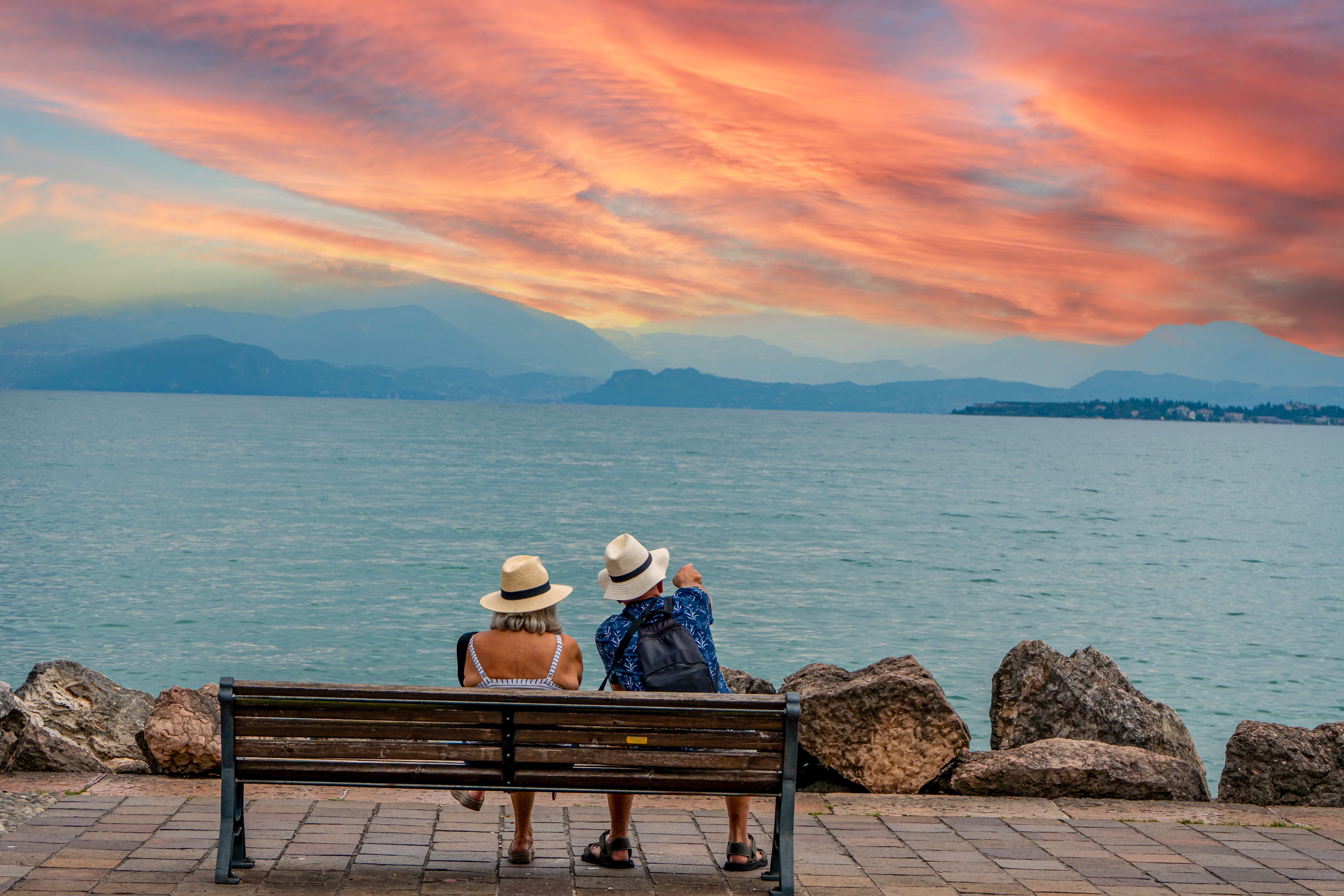 Ældre par nyder romantisk solnedgang på en bænk ved Gardasøen med udsigt til bjergene i Italien
