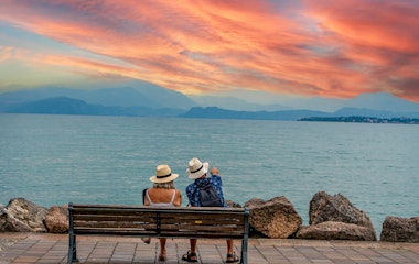 Ældre par nyder romantisk solnedgang på en bænk ved Gardasøen med udsigt til bjergene i Italien