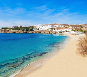 Idylliske Agios Stefanos strand på Mykonos med gyldent sand og krystalklart turkisblåt vand, omgivet af det karakteristiske kykladiske landskab under den græske sol