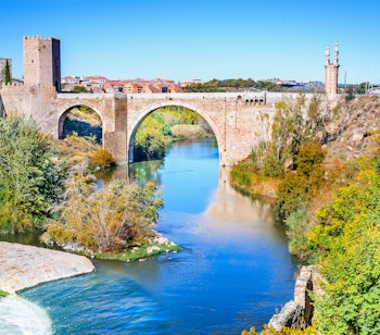 Den historiske Alcantara-bro med stenbuer over den blå Tagus-flod i Toledo, Spanien, omgivet af grøn vegetation og middelalderlige fæstningsværker
