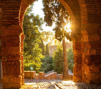Smuk solnedgang ved den historiske arabiske fæstning Alcazaba i Malaga med gyldent lys på de gamle stenmure og haver - et must-see i Andalusien