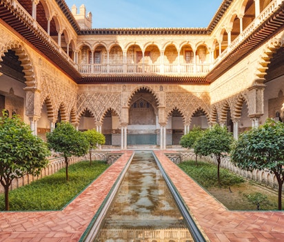 Smuk gårdhave i det kongelige Alcazar-palads i Sevilla med elegante søjler, mauriske buer og detaljerede dekorationer - oplev Andalusiens fascinerende blanding af islamisk og spansk arkitektur på din rejse til Spanien
