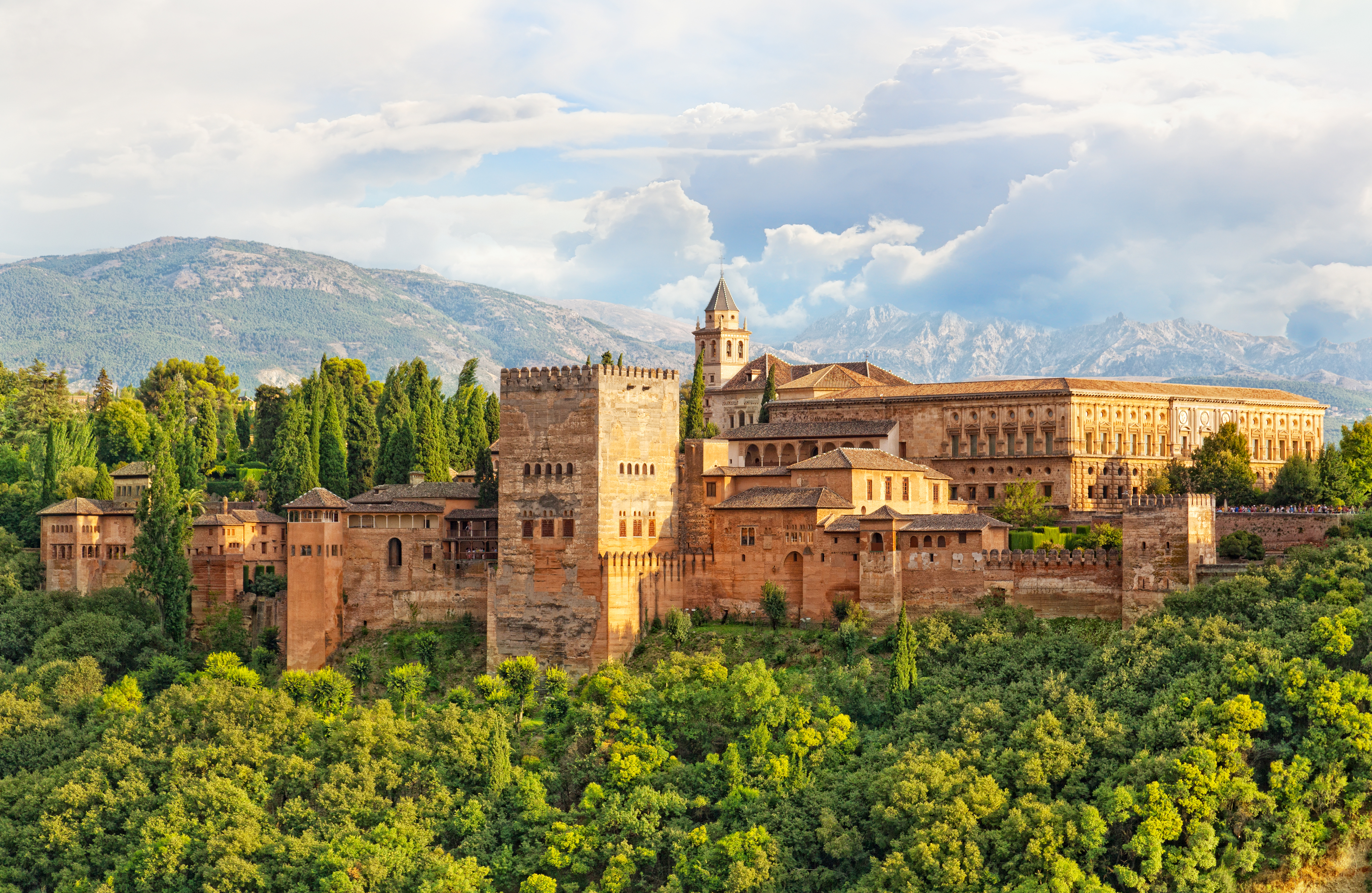 Den historiske Alhambra-fæstning i Granada, Spanien, med imponerende mauriske mure omgivet af grøn vegetation og med Sierra Nevada bjergene i baggrunden