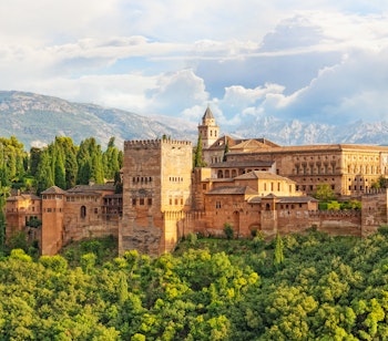 Den historiske Alhambra-fæstning i Granada, Spanien, med imponerende mauriske mure omgivet af grøn vegetation og med Sierra Nevada bjergene i baggrunden