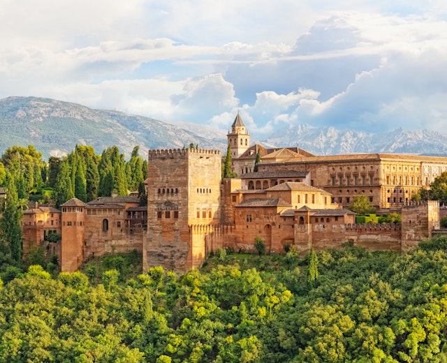 Den historiske Alhambra-fæstning i Granada, Spanien, med imponerende mauriske mure omgivet af grøn vegetation og med Sierra Nevada bjergene i baggrunden