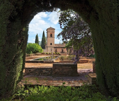 Udsigt gennem en grøn havebue til det historiske Alhambra palads og haver i Granada, Spanien med klokketårn og blå himmel