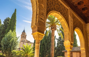 Historiske mauriske buer i Alhambra-paladset i Granada med udsigt til palmetræer og tårn i gyldent sollys, Andalusien, Spanien