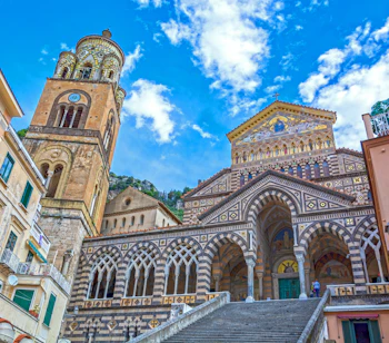 Den historiske domkirke i Amalfi med sin imponerende hvide facade og brede trappe på Piazza del Duomo under en klar blå sommerhimmel