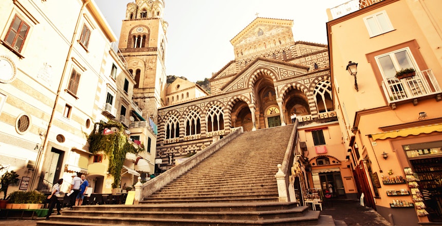 Den historiske domkirkeplads i Amalfi med den ikoniske Sankt Andreas Katedral og dens imponerende trappe og stribede byzantinsk-romanske facade, omgivet af traditionelle bygninger på Amalfikysten