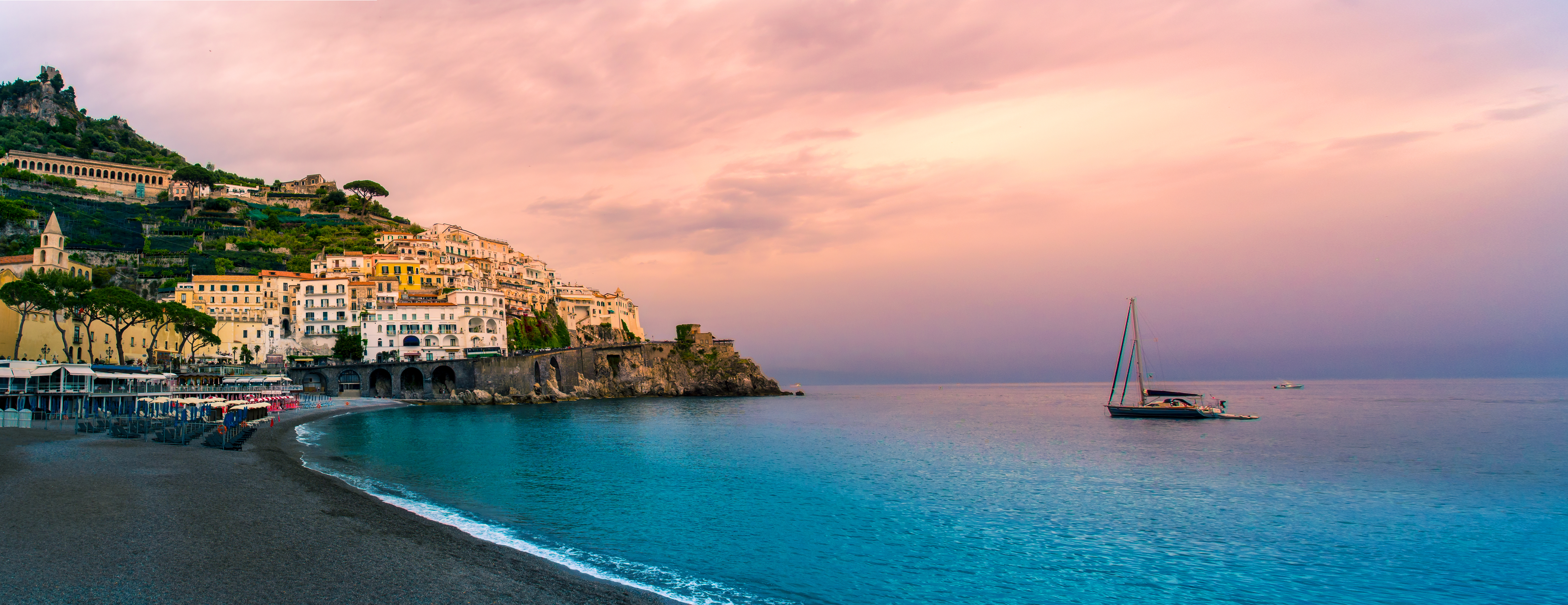 Betagende panoramaudsigt over Amalfikysten ved solnedgang med gyldne og rosa himmelfarver, der spejler sig i det turkisblå Middelhav og fremhæver den dramatiske kystlinje