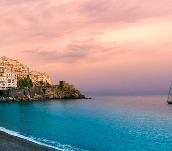 Betagende panoramaudsigt over Amalfikysten ved solnedgang med gyldne og rosa himmelfarver, der spejler sig i det turkisblå Middelhav og fremhæver den dramatiske kystlinje