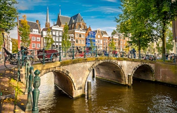 Idyllisk kanal med historisk stenbro ved Prinsengracht i Amsterdam, omgivet af farverige hollandske bygninger i solskin