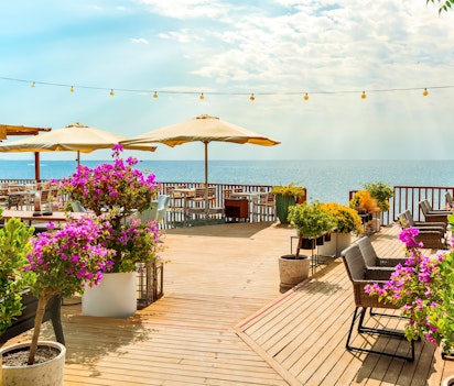 Idyllisk udendørs café i Antalya prydet med smukke røde blomster, hvor traditionelle borde og stole er placeret med udsigt over det turkisblå Middelhav, perfekt til en afslappende sommerferie