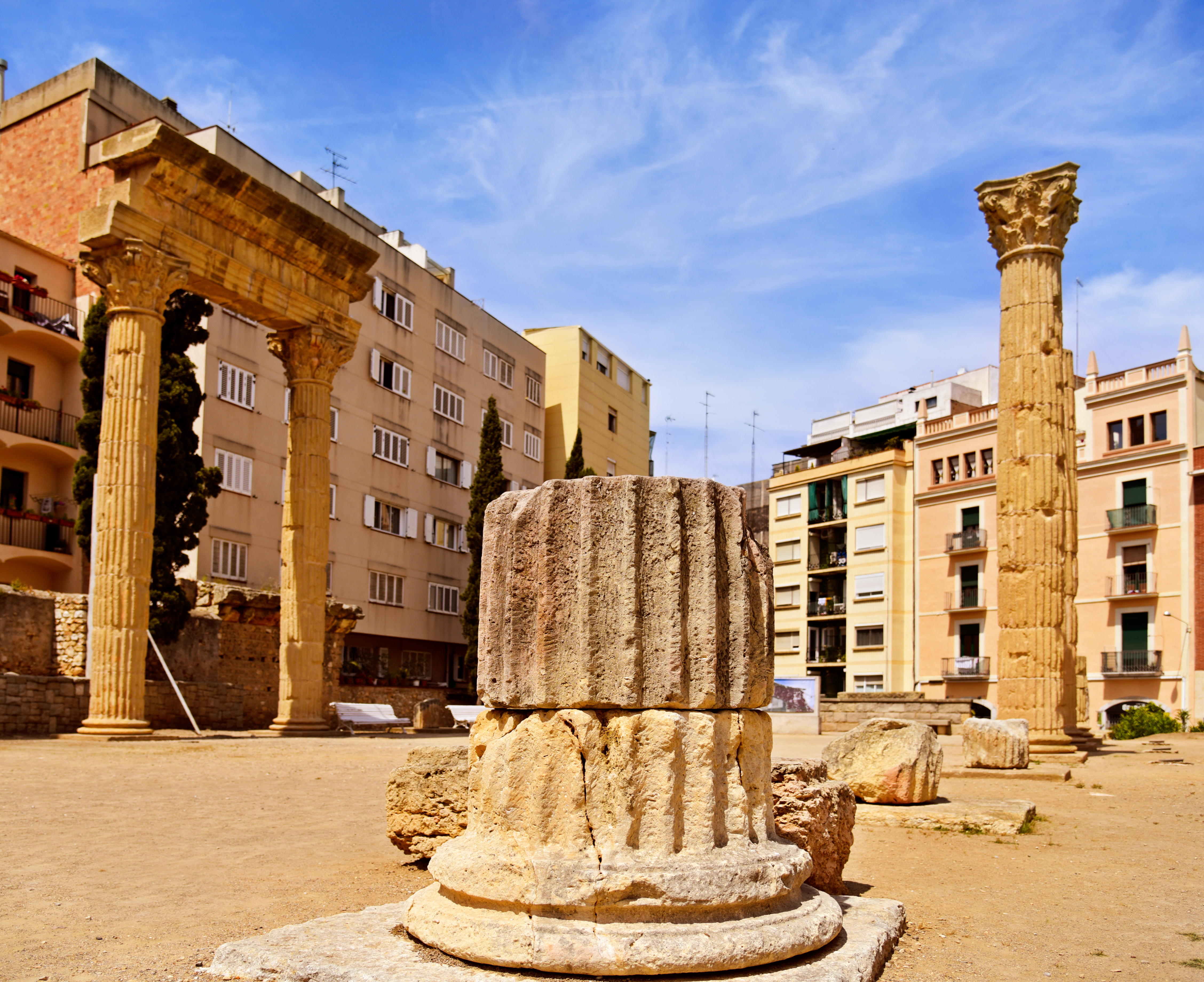 Antikke romerske tempelruiner med korinthiske søjler omgivet af moderne mediterrane bygninger under blå himmel