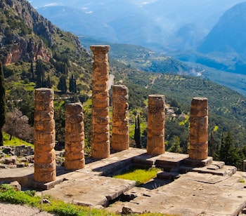 Fugleperspektiv af det antikke Apollon-tempel i Delfi, omgivet af fyrretræer med Parnassos-bjergene i baggrunden, Grækenland