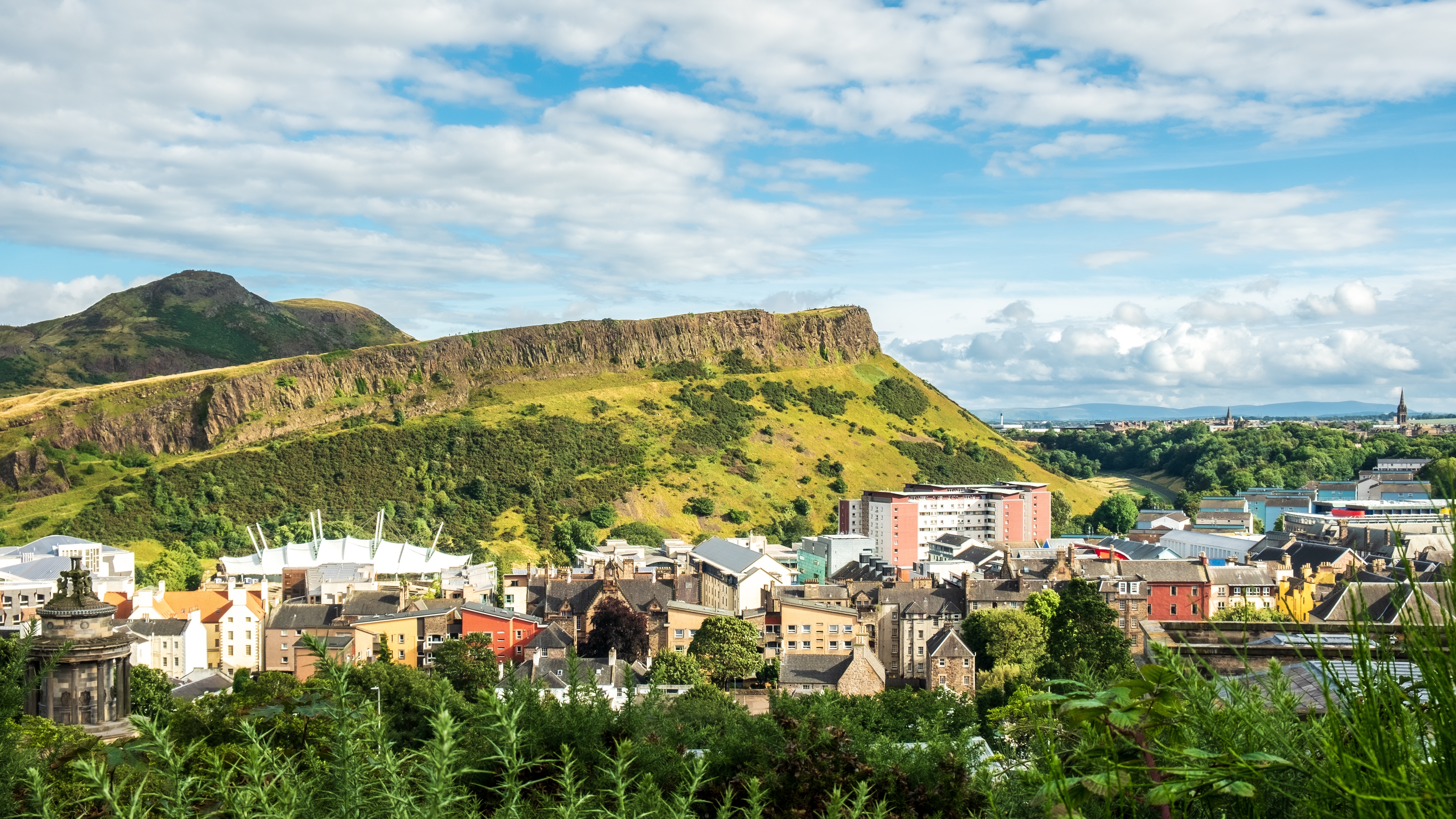 Panoramaudsigt over Arthur's Seat bjerget i Edinburgh med grønne naturområder og byens skyline under blå himmel