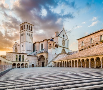 Den historiske Basilika San Francesco i Assisi badet i gyldent solnedgangslys med sit ikoniske klokketårn og søjlegang i Umbrien, Italien - et kulturelt højdepunkt på elcykelturen gennem det centrale Italien