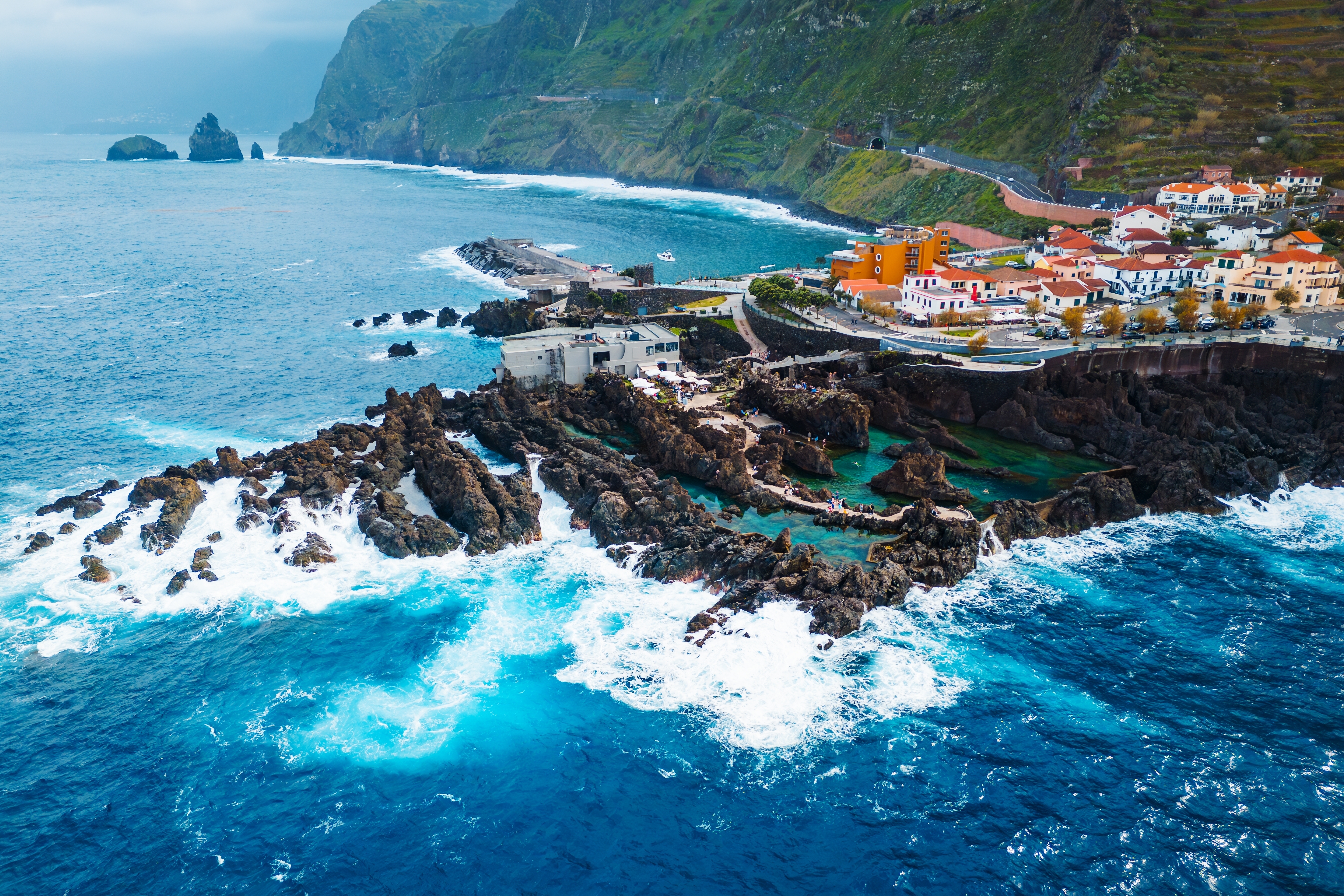 Dramatisk kystlandsby med vulkanske klipper og turkisblå oceanbølger der bryder mod sorte klippefomationer