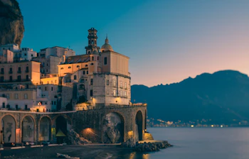 Aftenstemning over Atrani på Amalfikysten med den oplyste katedral, historiske stenbuer ud mod havet og bjerge i baggrunden