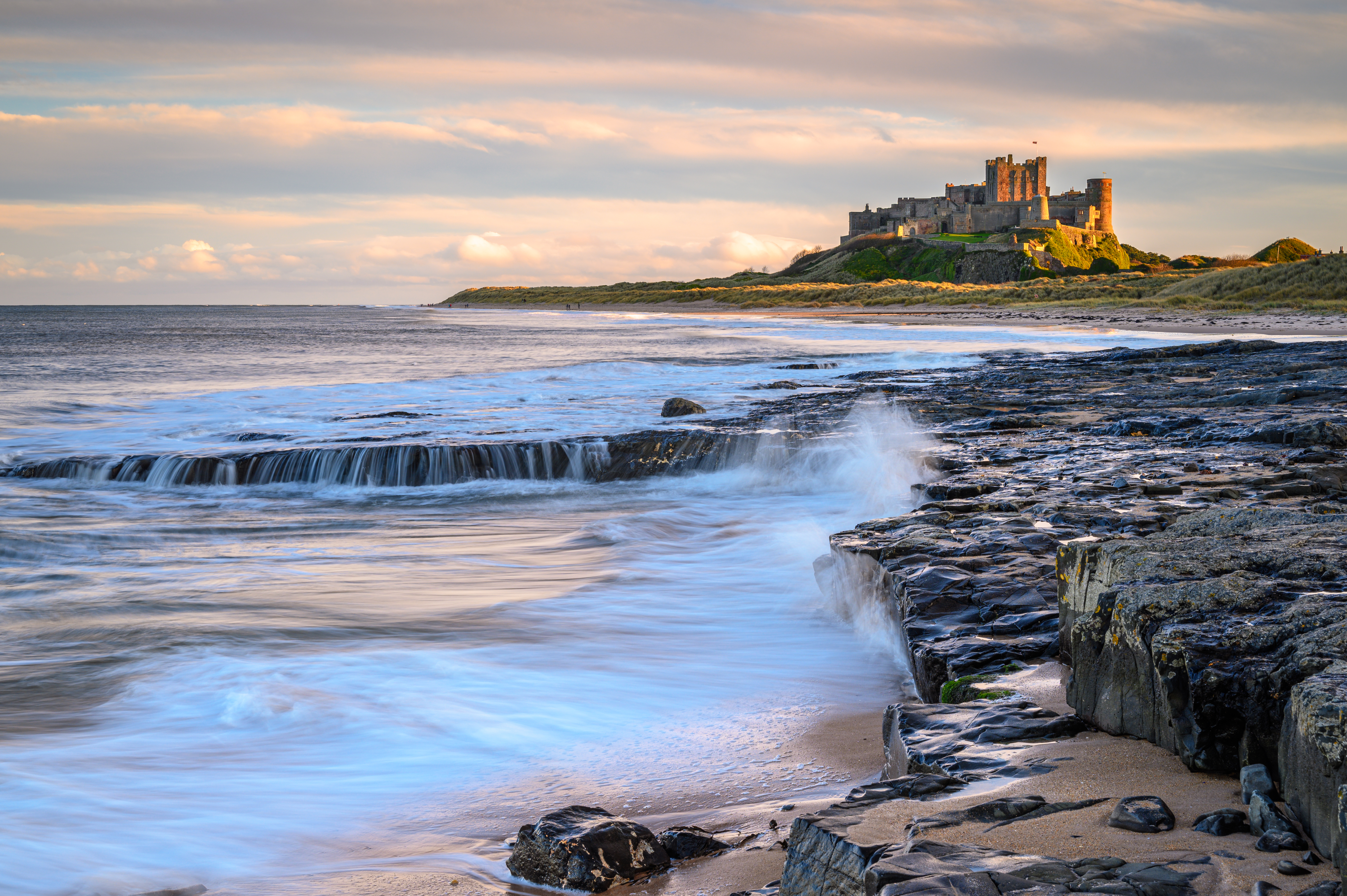 Bamburgh Castle tronende over Northumberlands klippekyst med bølger der skyller ind over de mørke klipper ved solnedgang