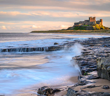 Bamburgh Castle tronende over Northumberlands klippekyst med bølger der skyller ind over de mørke klipper ved solnedgang