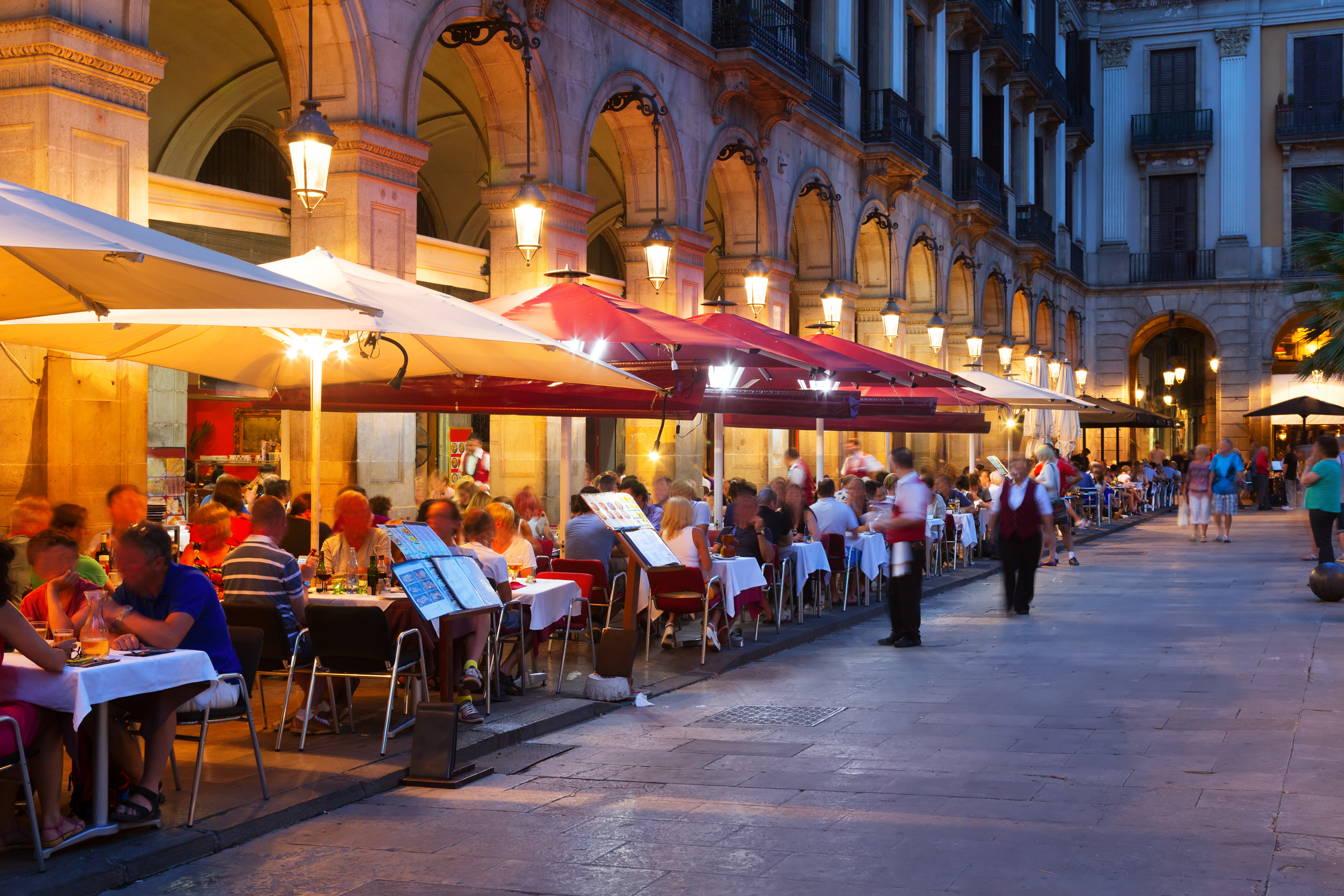 Stemningsfuld sommeraften på Placa Reial i Barcelona med oplyste udeserveringer, historiske arkader og besøgende, der nyder det livlige catalanske natteliv på den ikoniske plads