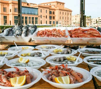 Farverigt udendørs fiskemarked på Baris promenade i Apulien, hvor friske havfrugter som rejer, østers, blæksprutter og fisk præsenteres med citronskiver klar til at blive nydt på stedet