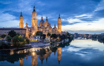 Stemningsfuld aftenvy over den oplyste Basilica del Pilar i Zaragoza, hvis imponerende barokarkitektur og karakteristiske tårne spejler sig perfekt i Ebro-flodens rolige vand