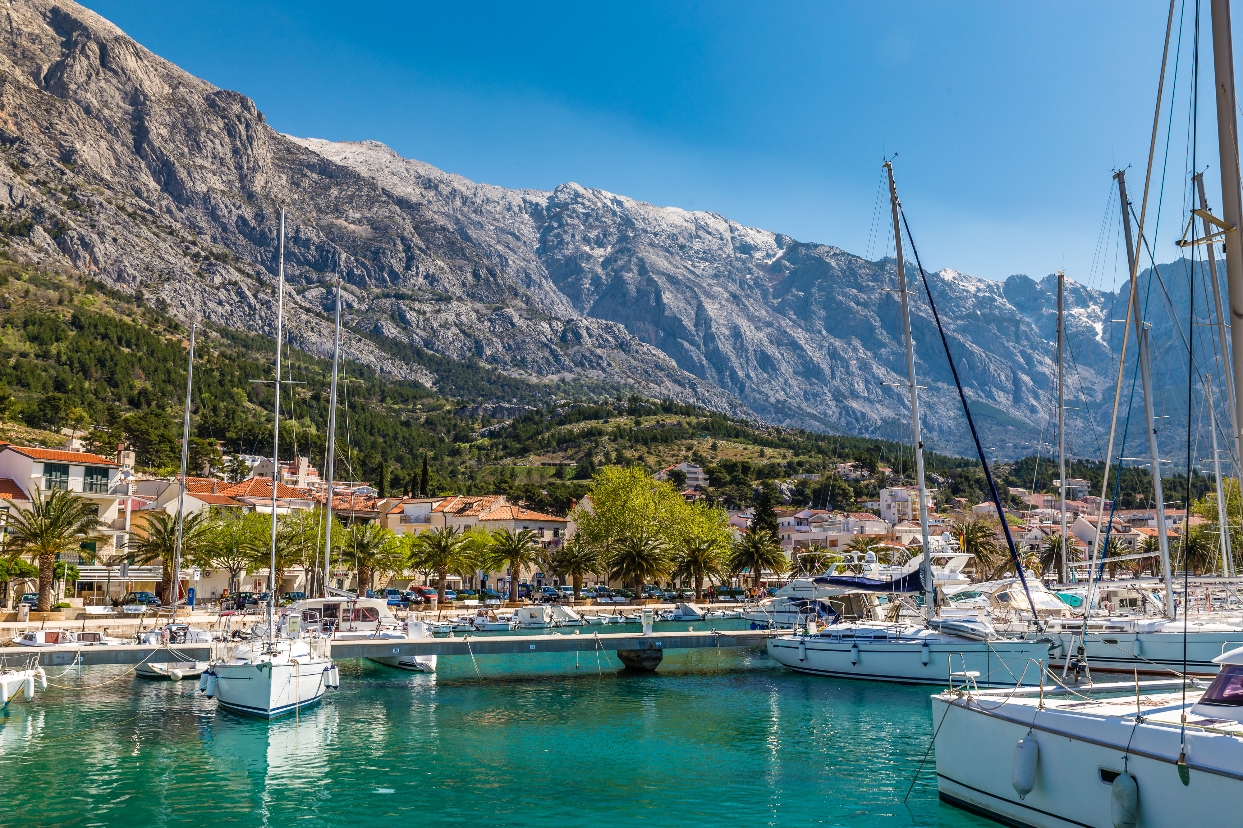 Idyllisk havnefront i Baska Voda med sejlbåde, farverige bygninger og det majestætiske Biokovo-bjerg i baggrunden, Makarska Riviera, Dalmatien, Kroatien