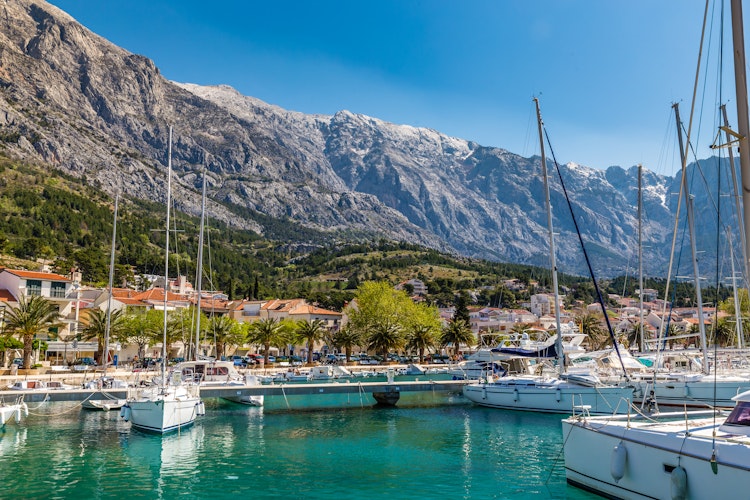 Idyllisk havnefront i Baska Voda med sejlbåde, farverige bygninger og det majestætiske Biokovo-bjerg i baggrunden, Makarska Riviera, Dalmatien, Kroatien