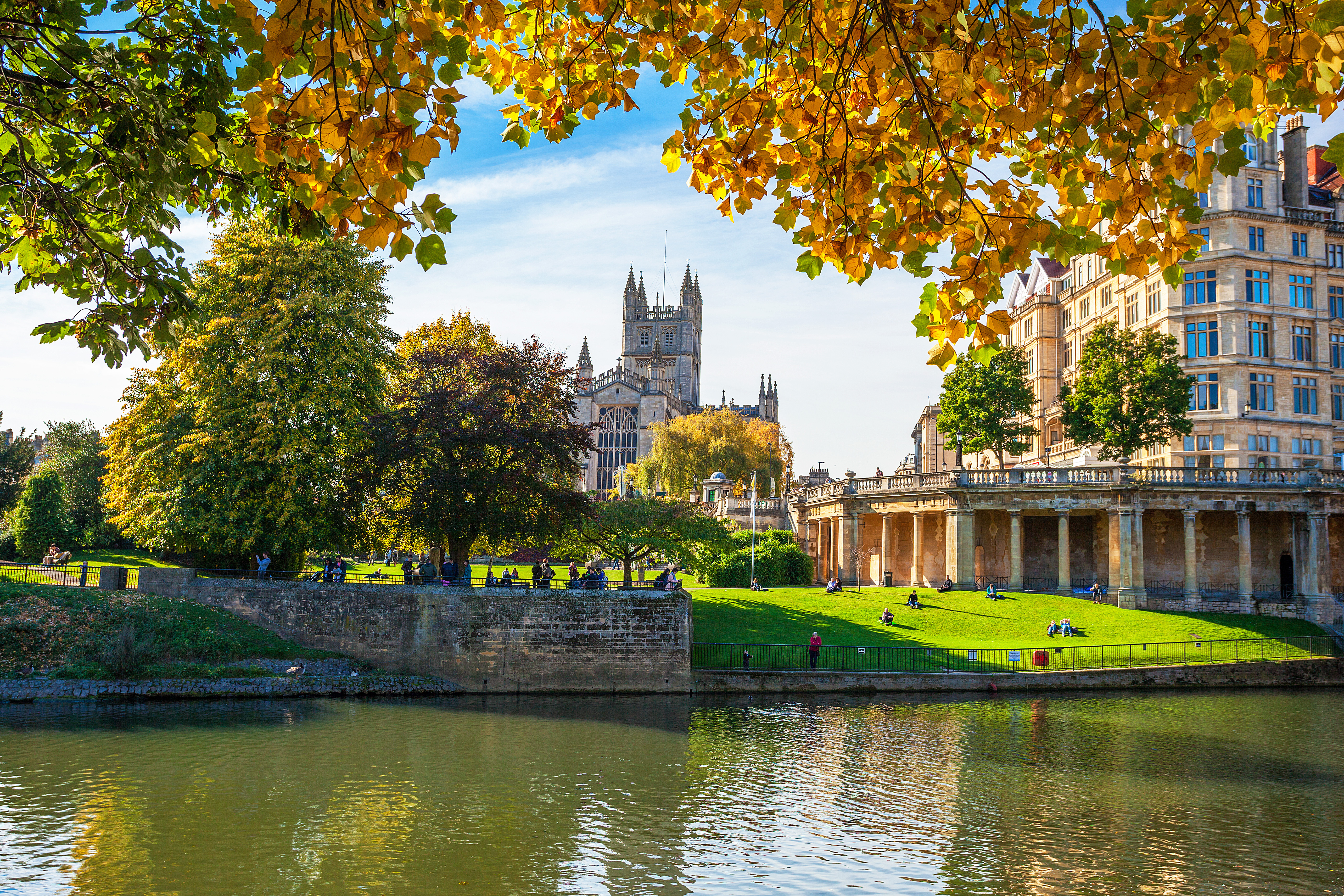 Bath Abbey og floden Avon i efterårsfarver med gyldne træer, der indrammer den historiske katedral og flodpromenaden i Bath, England