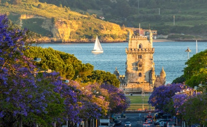 Det historiske Belem-tårn omgivet af lilla jacaranda-træer ved Tagus-floden under solnedgang i Lissabon, Portugal