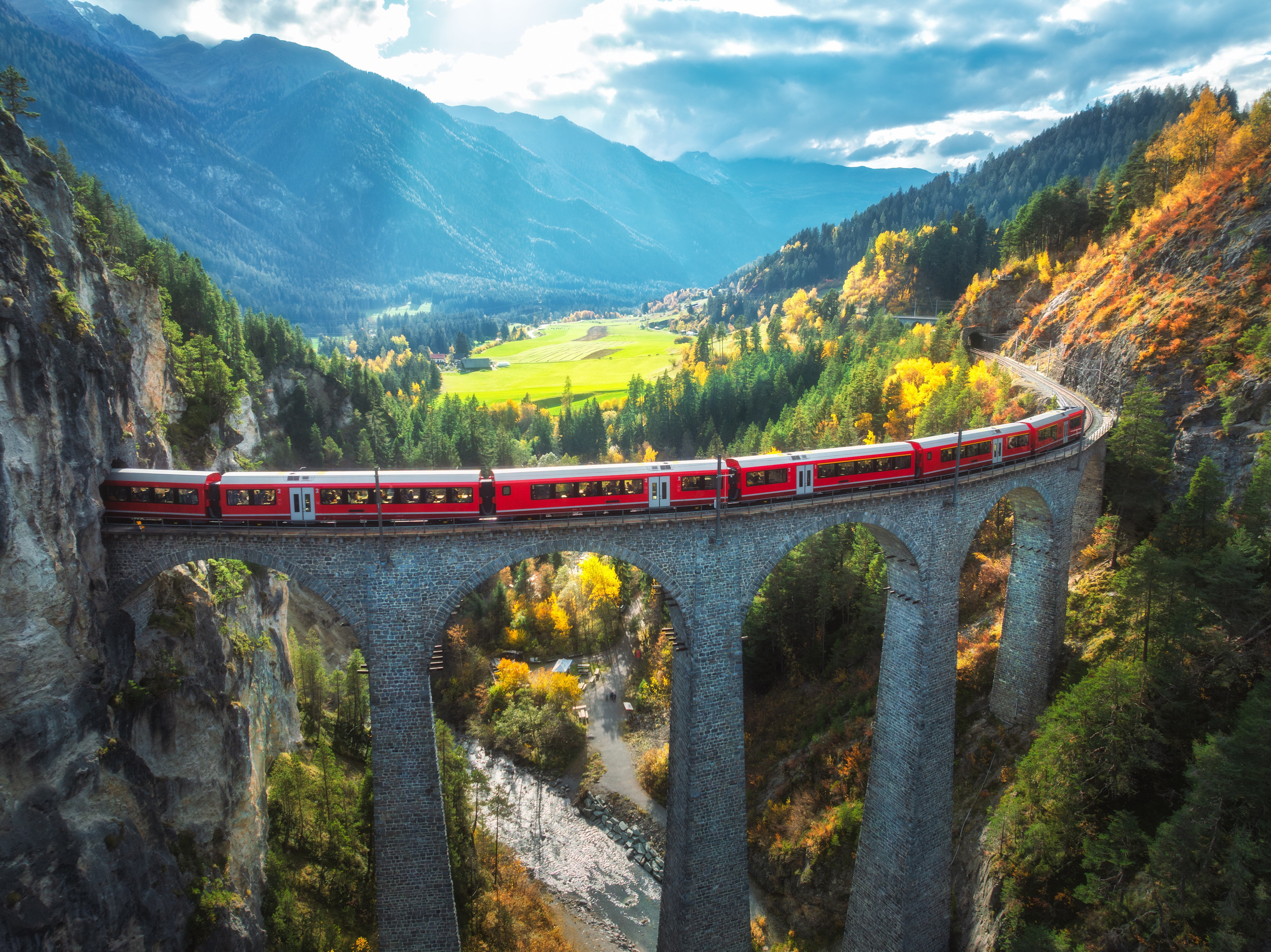 Luftfoto af det røde Bernina Express tog der krydser den berømte Landwasser Viadukt i de schweiziske alper, omgivet af farverige efterårsskove ved solnedgang
