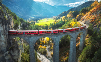 Luftfoto af det røde Bernina Express tog der krydser den berømte Landwasser Viadukt i de schweiziske alper, omgivet af farverige efterårsskove ved solnedgang