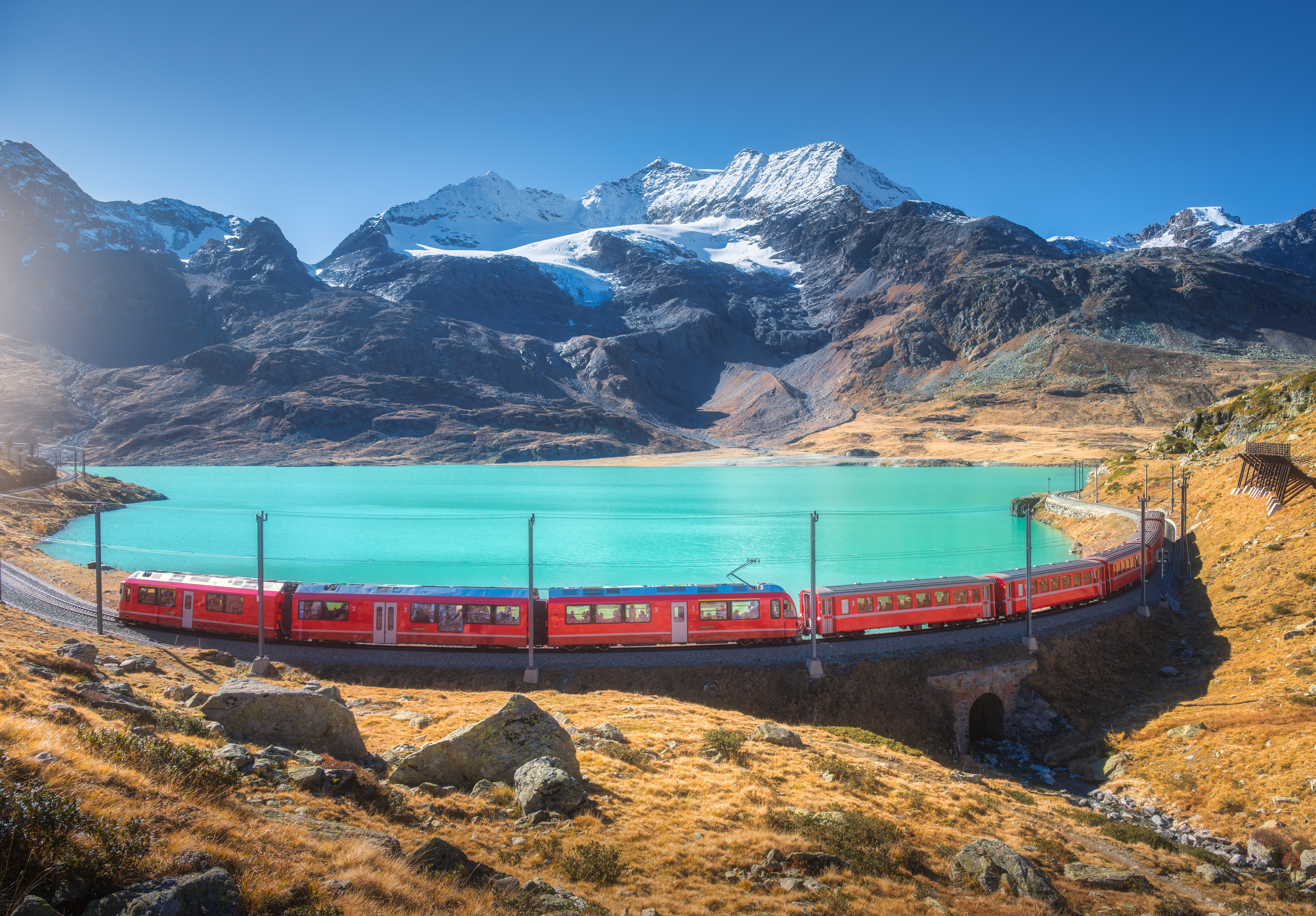Rødt Bernina Express tog kører gennem schweiziske alper ved turkisblå alpesø