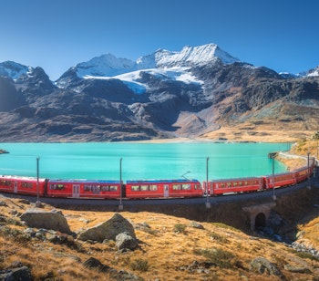 Rødt Bernina Express tog kører gennem schweiziske alper ved turkisblå alpesø