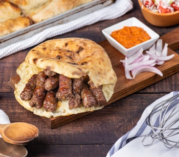 Traditionel bosnisk čevapi serveret med somun-brød, hakket løg og krydderier på træskærebræt - en autentisk smagsoplevelse fra Balkan
