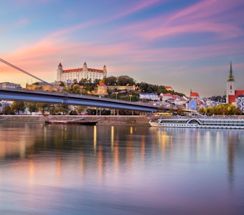 Malerisk solnedgang over Bratislava med det historiske slot på bakketoppen, Donaufloden og byens charmerende skyline badet i gyldne og lilla efterårsfarver.