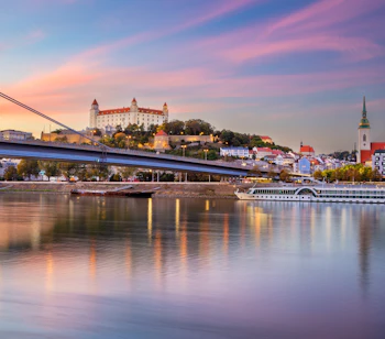 Malerisk solnedgang over Bratislava med det historiske slot på bakketoppen, Donaufloden og byens charmerende skyline badet i gyldne og lilla efterårsfarver.