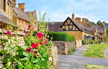 Idyllisk gade i Broadway landsbyen i Cotswolds med smukke røde stokroser langs stenmure og traditionelle honningfarvede stenhuse under en sommerblå himmel