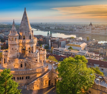 Gylden solopgang ved Fiskerbastionen i Budapest med udsigt til parlamentsbygningen og Donau-floden under blå sommerhimmel