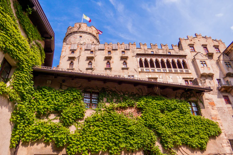 Buonconsiglio Slot i Trento, Italien, med efeudækkede mure, middelalderlige tinder og italiensk flag på tårnet mod blå himmel