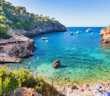 Den idylliske bugt Cala Deia på Mallorca med krystalklart azurblåt vand, dramatiske klipper og små fiskerbåde - en skjult perle ved Middelhavets kyst