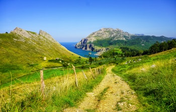 Naturskøn grusvej langs Camino de Santiago i Cantabria med frodig grøn natur og betagende udsigt over Atlanterhavet under en klar blå himmel i Nordspanien