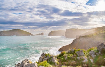 Betagende kystlandskab langs Camino Norte nær Llanes i Asturien, med dramatiske klipper, turkisblå Atlanterhavsbølger og gylden morgenlys over den barske kystlinje