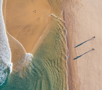 Luftfoto af et par der vandrer langs en smuk sandstrand ved Atlanterhavet under solopgang på Camino Portugués ruten, med lange skygger på sandet