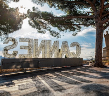 Historisk udsigt over Cannes med Notre-Dame kirken, World Exploration Museum skilt og Vor Frue statue omgivet af solbeskinnede pinjetræer