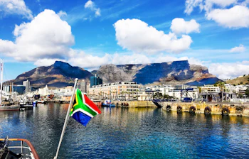 Smuk udsigt over Cape Town Waterfront med Table Mountain i baggrunden og både i Victoria & Alfred havnen under blå himmel med skyer og sydafrikansk flag