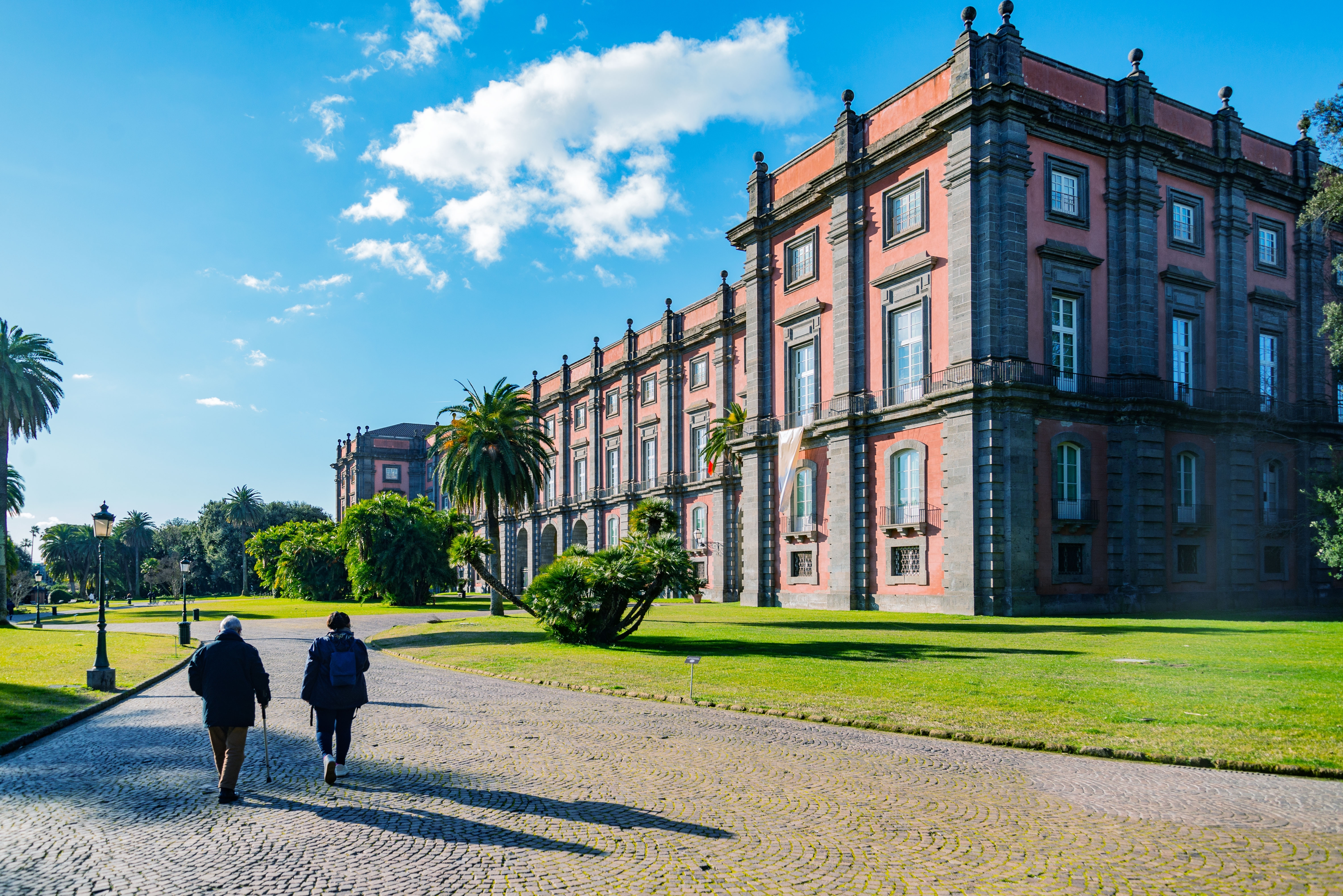 To besøgende går ad en brolagt sti mod det majestætiske Capodimonte-palads og museum i Napoli, Italien, omgivet af grønne haver og palmer under en strålende blå himmel